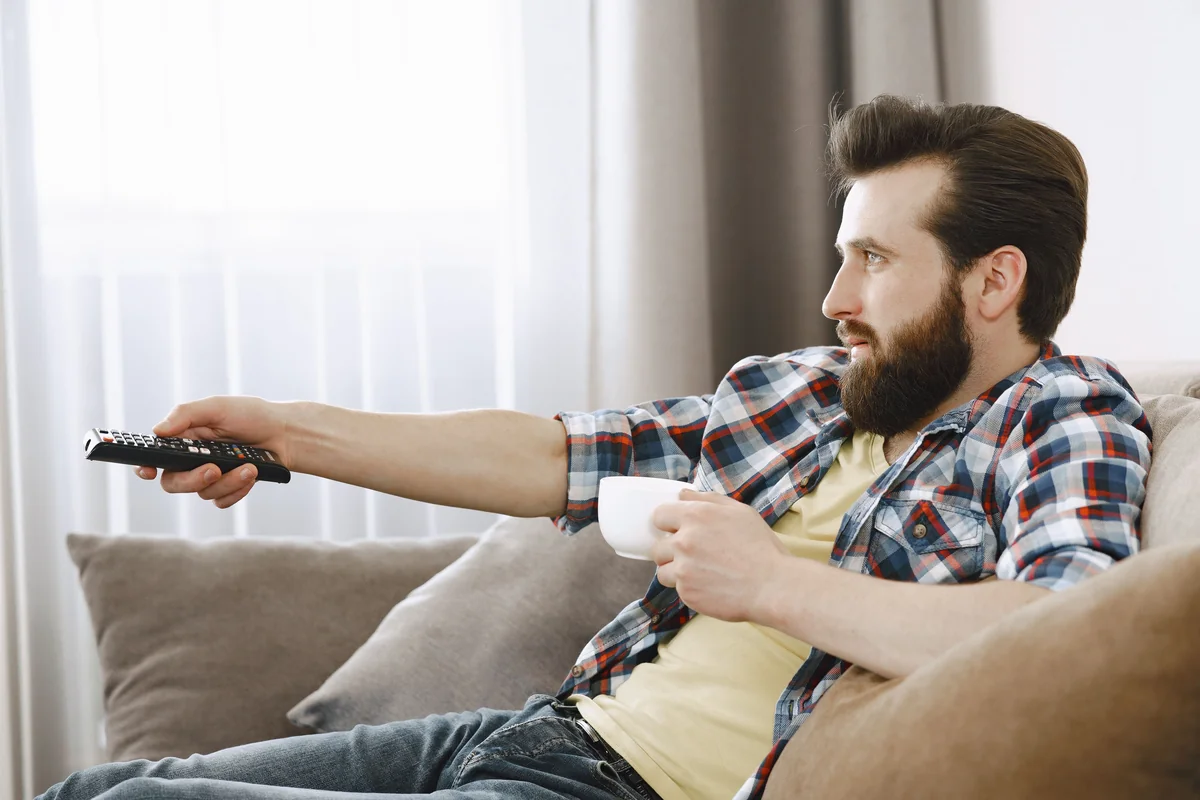 Person sitzt auf einer Couch und richtet eine Fernbedienung auf einen Fernseher