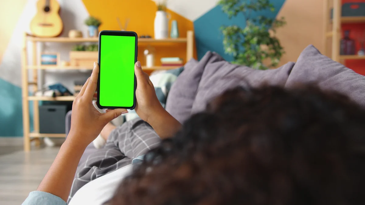Person hält ein Smartphone, während im Hintergrund ein Fernseher im Wohnzimmer läuft